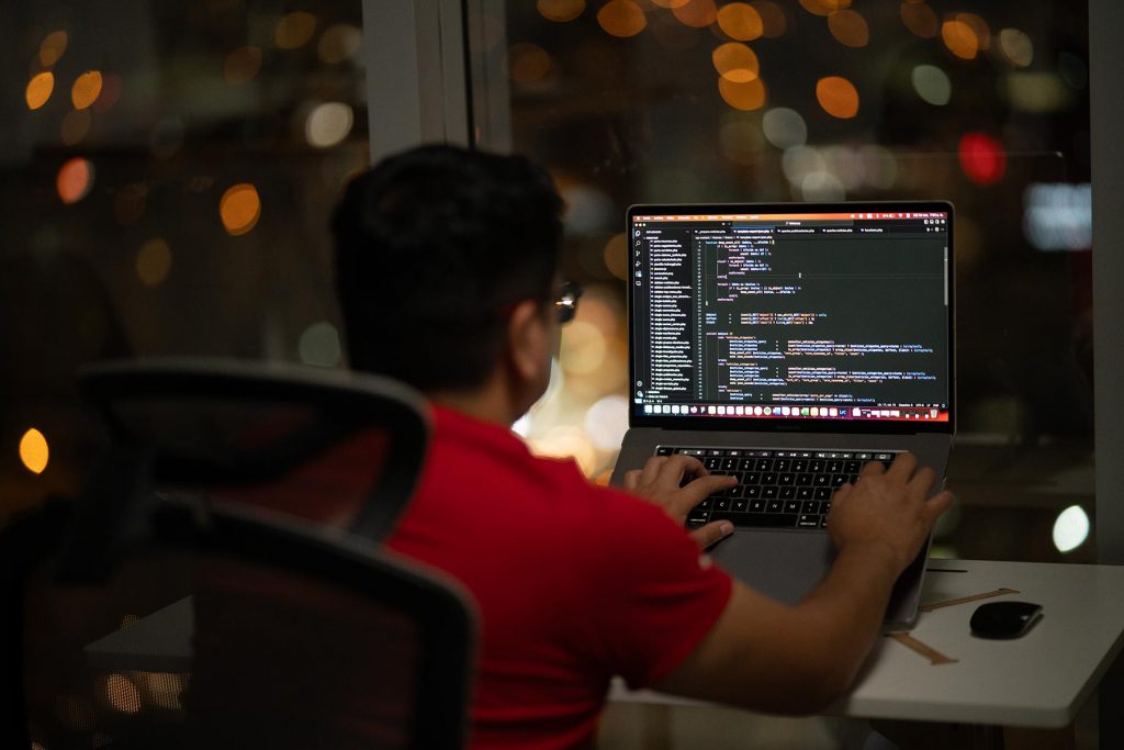 Hombre trabajando en el desarrollo web de un proyecto con una laptop, frente a una ventana por la noche.
